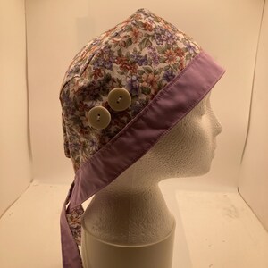 May include: A scrub cap featuring a floral pattern with orange, purple, and green flowers on a white background. The cap has a solid lavender band along the bottom edge and two round, light-coloured buttons attached to the side for mask support.