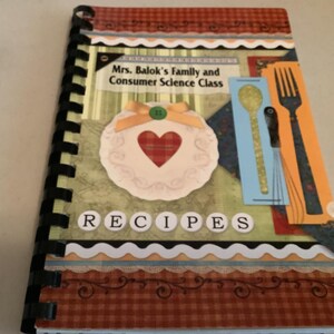Op de afbeelding: Een receptenboek met spiraalbinding en een decoratieve omslag. De omslag toont de tekst "Mrs. Balok's Family and Consumer Science Class" en "RECIPES". Het bevat een hartvormig ontwerp, een lepel en een vorkillustratie.