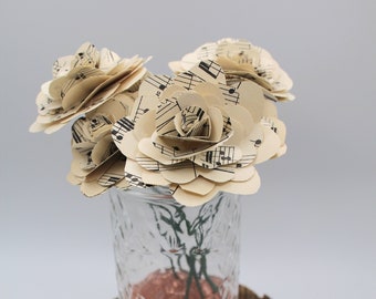May include: A bouquet of paper roses made from sheet music is arranged in a clear glass jar with copper glitter. The jar is sitting on a wooden slice.