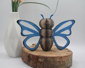 Puede incluir: Una escultura de mariposa azul hecha de páginas de libros recicladas. La mariposa está sentada sobre una base de madera.