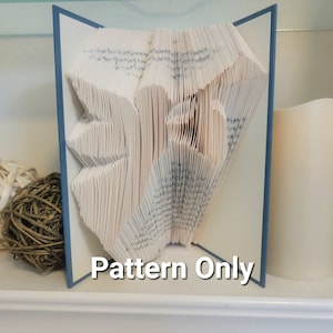 May include: A book folded into the shape of a heart, with a blue cover and the words "Pattern Only" printed on the bottom. The book is sitting on a white shelf.