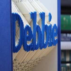 Puede incluir: Un libro con el nombre "Debbie" doblado en las p&aacute;ginas. El libro es azul y las letras son azules. El libro est&aacute; en un estante con otros libros.