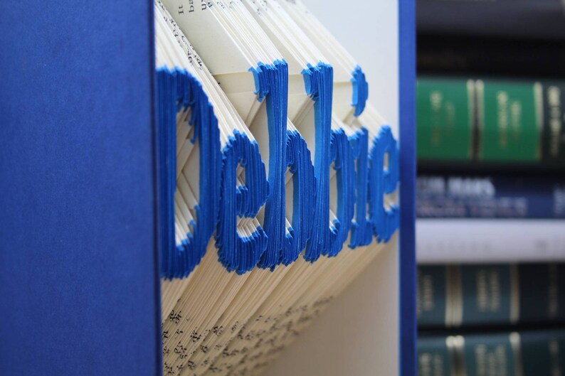 Puede incluir: Un libro con el nombre "Debbie" doblado en las p&aacute;ginas. El libro es azul y las letras son azules. El libro est&aacute; en un estante con otros libros.