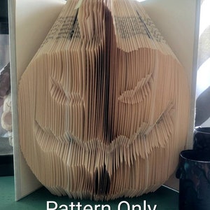 May include: A book folded into the shape of a jack-o'-lantern. The book is made of beige paper and has a white cover. The text "Pattern Only" is printed in black at the bottom of the image.