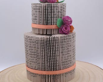 Puede incluir: Pastel de libro de dos niveles decorado con flores de papel rosa y naranja. El pastel está hecho de páginas de un libro y está atado con una cinta de color melocotón.