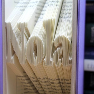 Puede incluir: Un libro con el nombre "Nolan" plegado en las p&aacute;ginas. El libro est&aacute; encuadernado en morado y est&aacute; en una estanter&iacute;a con otros libros.