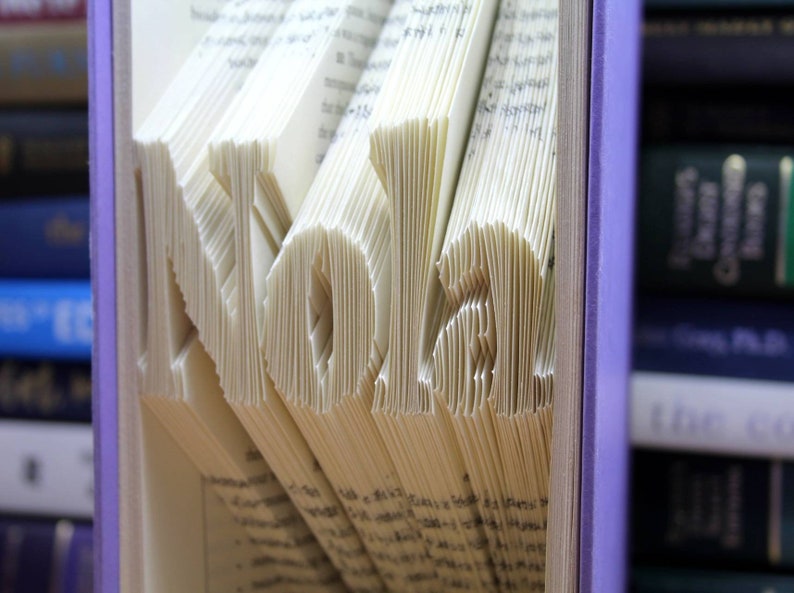 Puede incluir: Un libro con el nombre "Nolan" plegado en las p&aacute;ginas. El libro est&aacute; encuadernado en morado y est&aacute; en una estanter&iacute;a con otros libros.