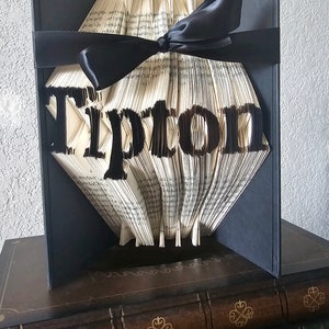 Puede incluir: Una escultura de arte de libro con la palabra "Tipton" plegada en las p&aacute;ginas. El libro est&aacute; encuadernado en tela negra y atado con una cinta negra. La escultura se exhibe sobre una pila de libros.
