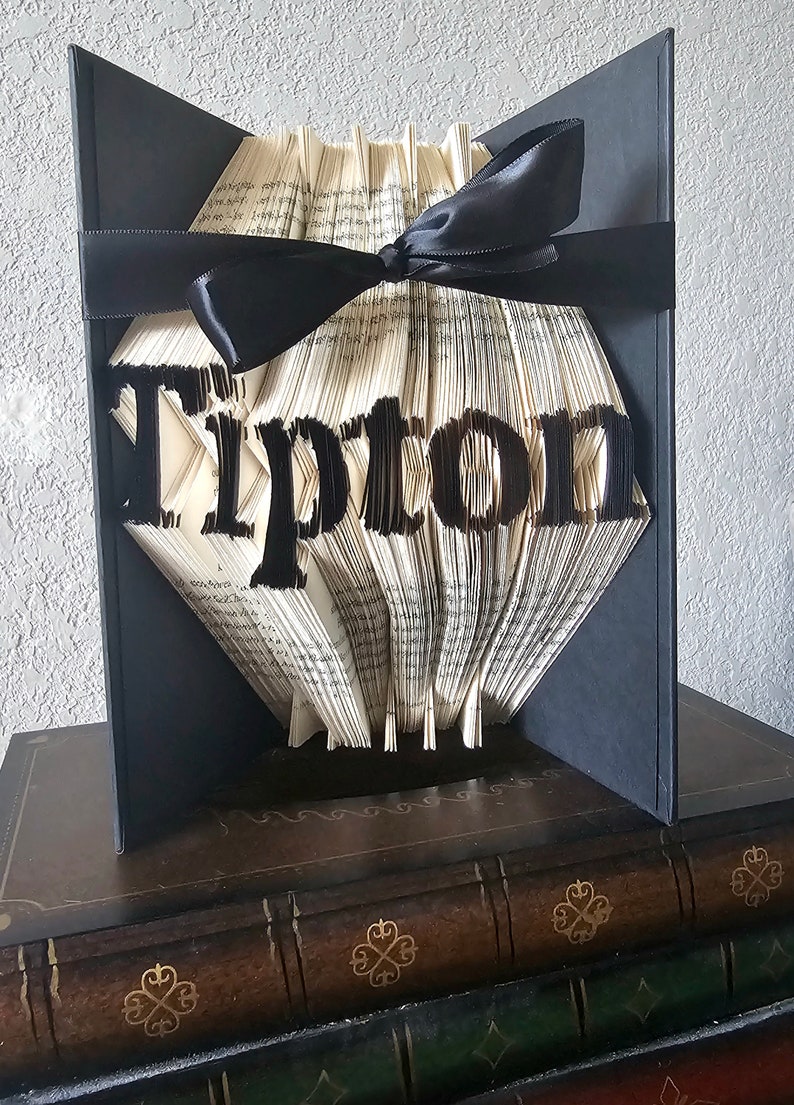 Puede incluir: Una escultura de arte de libro con la palabra "Tipton" plegada en las p&aacute;ginas. El libro est&aacute; encuadernado en tela negra y atado con una cinta negra. La escultura se exhibe sobre una pila de libros.
