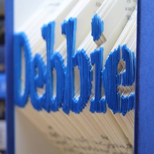 Puede incluir: Un libro con el nombre "Debbie" escrito en papel azul en la portada. El libro est&aacute; cerrado y la columna vertebral es visible.