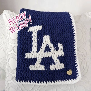May include: A blue and white crocheted blanket with the letter "LA" in white yarn. The blanket has a white border and a small heart-shaped tag in the bottom right corner.