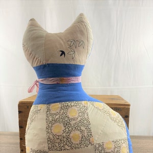 May include: A blue and white patchwork cat pillow with a pink ribbon around its neck. The cat has a simple embroidered design on its back.
