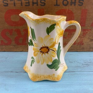 May include: A white ceramic pitcher with a yellow and green floral design. The pitcher has a scalloped rim and a handle.