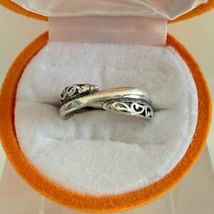 May include: A silver-toned ring featuring a crossover design with intricate cut-out scrollwork patterns on the overlapping ends. The band has a polished finish and is displayed inside a round, bright orange velvet-lined jewelry box.