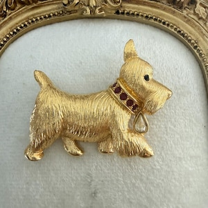 May include: A gold-toned metal brooch in the shape of a Scottish Terrier dog. The dog has a textured coat, a red collar with small stones, and a teardrop-shaped charm. The brooch is set against a white background.