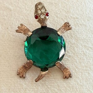 May include: A vintage turtle brooch with a large, faceted emerald green gemstone body. The turtle's head is adorned with clear and red rhinestones, and the legs and head are gold-toned metal. The brooch is a unique piece of jewelry.