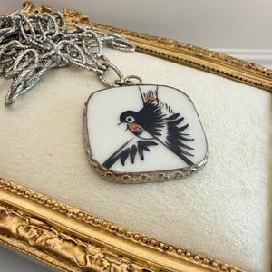 May include: A silver chain necklace with a square pendant featuring a bird illustration. The pendant has a white background with a black and red bird design. The pendant is framed with a silver border.
