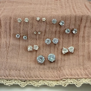 May include: A collection of various silver-toned stud earrings featuring clear, faceted stones in multiple shapes including round, star, and heart designs. The set includes several pairs of earrings with different sizes and stone cuts.