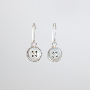 May include: A pair of silver button earrings with a white button design. The buttons have two holes.