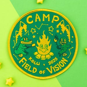 Pode incluir: Patch bordado com um design verde e amarelo. O patch apresenta o texto "CAMP FIELD OF VISION 2025" e ilustrações de uma fogueira, um crocodilo e um pato. Enfeites amarelos em forma de estrela estão espalhados ao redor do patch.