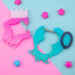 Peut inclure: Deux porte-clés fantaisistes, roses et turquoise, chacun avec un fermoir argenté. Le porte-clés rose a un motif de couronne, tandis que celui turquoise a une forme unique. Des étoiles roses et bleues sont éparpillées. Le texte "US TARIFFS INCLUDED!" est visible.
