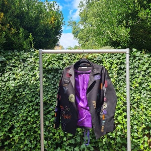 Może przedstawiać: Czarna kurtka z kolorowymi haftowanymi naszywkami i tekstem, w tym "Disco Cool" i "Hey So Light". Kurtka ma fioletową podszewkę i wisi na metalowym wieszaku na tle zielonych liści i błękitnego nieba.