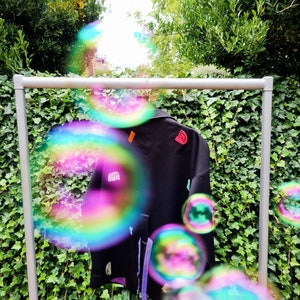 Może przedstawiać: Czarna kurtka z kolorowymi naszywkami i fioletową podszewką wisi na metalowym stojaku. Kurtka ma kołnierz i kieszenie. Naszywki obejmują tęczę, literę "D" i inne abstrakcyjne kształty. Tło to zielona ściana z bluszczu.