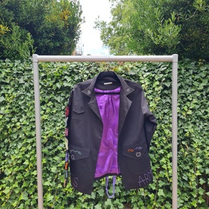 Może przedstawiać: Czarna kurtka z fioletową podszewką, wisząca na metalowym stojaku. Kurtka ma haftowane detale, w tym słowa "na-na" i "ARMS" oraz kolorowe naszywki. Posiada dwie przednie kieszenie i klasyczny kołnierz.