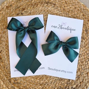 May include: Two dark green satin hair bows on white cards, one with long streamers. The cards have the text "one2boutique" and "2boutique.etsy.com". The bows are displayed on a woven, tan surface.