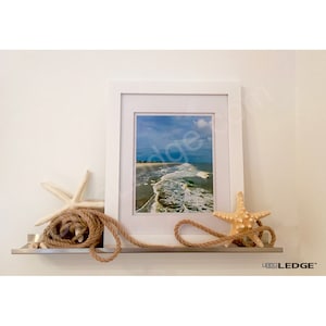 May include: A white framed print of a beach scene, displayed on a silver shelf. Decorative elements include a white starfish, a tan starfish, and coiled rope. The shelf is mounted on a white wall.