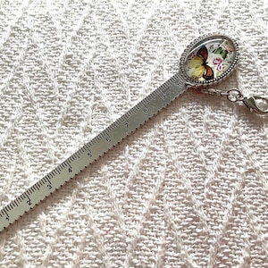 May include: A silver-toned metal bookmark ruler featuring a printed oval cabochon at the top with a butterfly and floral design. The ruler is marked with numbers from 1 to 10, indicating a length of 10 centimeters, and includes a lobster clasp.
