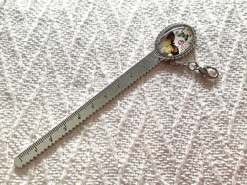 May include: A silver-toned metal bookmark ruler featuring a printed oval cabochon at the top with a butterfly and floral design. The ruler is marked with numbers from 1 to 10, indicating a length of 10 centimeters, and includes a lobster clasp.