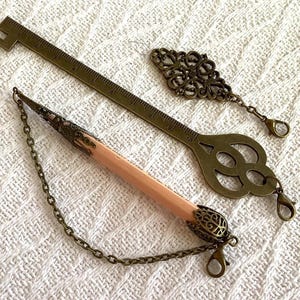 Pode incluir: Uma variedade de artigos de papelaria em tom de lat&atilde;o antigo. Inclui uma r&eacute;gua de 25 cm, um marcador decorativo em forma de chave, um berloque de filigrana e um l&aacute;pis com extremidade decorativa e corrente. Os itens est&atilde;o dispostos em uma superf&iacute;cie branca texturizada.
