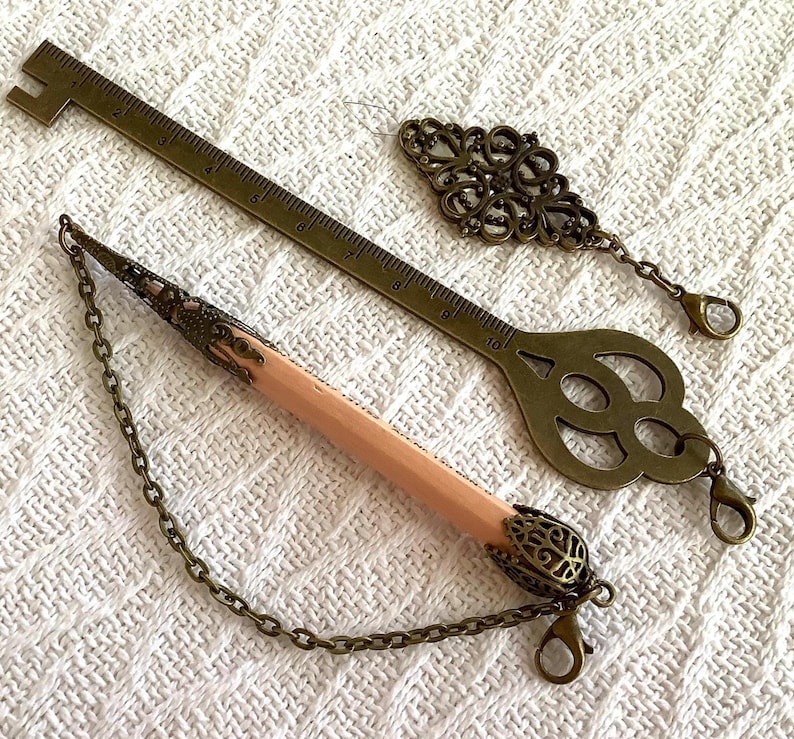 Pode incluir: Uma variedade de artigos de papelaria em tom de lat&atilde;o antigo. Inclui uma r&eacute;gua de 25 cm, um marcador decorativo em forma de chave, um berloque de filigrana e um l&aacute;pis com extremidade decorativa e corrente. Os itens est&atilde;o dispostos em uma superf&iacute;cie branca texturizada.