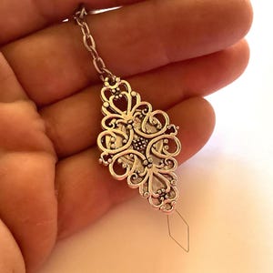 May include: A silver-toned metal pendant featuring an intricate, openwork filigree design with symmetrical scroll patterns. The pendant is attached to a short silver chain and has a thin, diamond-shaped wire loop extending from the bottom.