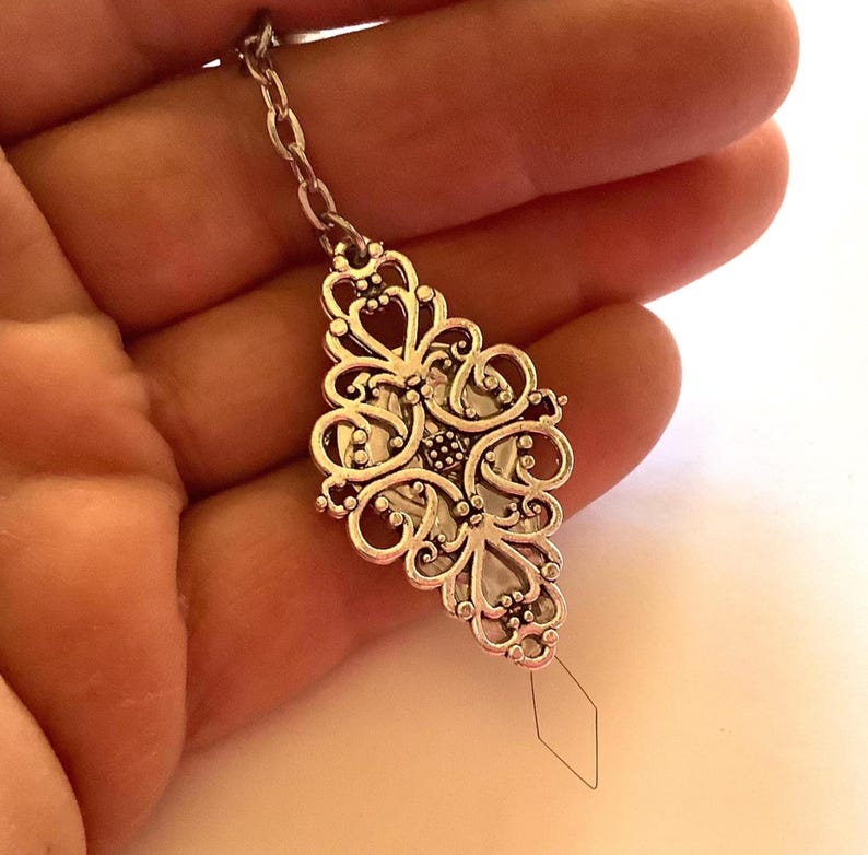 May include: A silver-toned metal pendant featuring an intricate, openwork filigree design with symmetrical scroll patterns. The pendant is attached to a short silver chain and has a thin, diamond-shaped wire loop extending from the bottom.