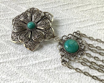 May include: An elaborate silver-coloured brooch in the shape of a flower, with a turquoise centre stone. It's linked to a smaller pendant with a turquoise stone and multiple chain dangles. The design has detailed filigree patterns.