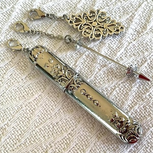 May include: Silver-toned needle case with floral and scrollwork details, a decorative charm, and a needle with a red tip. The case has a hinged lid and a chain with a clasp. The charm is an ornate oval shape.