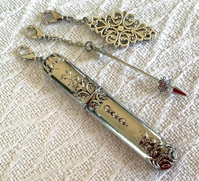 May include: Silver-toned needle case with floral and scrollwork details, a decorative charm, and a needle with a red tip. The case has a hinged lid and a chain with a clasp. The charm is an ornate oval shape.