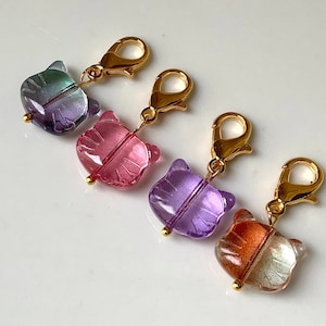May include: Four colourful glass cat-shaped stitch markers with gold metal clasps. The markers are purple, pink, blue and orange.