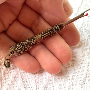 May include: A vintage-style, ornate, gold-toned threader with a red tip. The threader has a decorative floral design and a loop at the end.