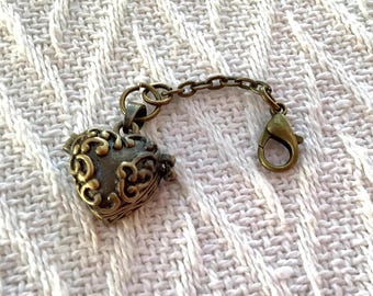 May include: A bronze-colored heart-shaped locket with ornate scrollwork, attached to a chain and clasp. The locket has a hinged design and a vintage aesthetic. The chain and clasp are also bronze-toned, creating a cohesive look.