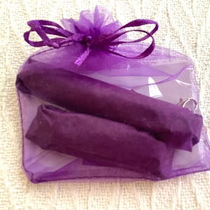 Pode incluir: Uma pequena sacola de presente roxa transparente amarrada com uma fita combinando. Dentro, dois itens enrolados s&atilde;o vis&iacute;veis, embrulhados em papel roxo escuro. A sacola est&aacute; em uma superf&iacute;cie texturizada esbranqui&ccedil;ada.