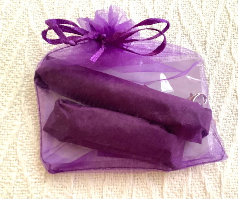 Pode incluir: Uma pequena sacola de presente roxa transparente amarrada com uma fita combinando. Dentro, dois itens enrolados s&atilde;o vis&iacute;veis, embrulhados em papel roxo escuro. A sacola est&aacute; em uma superf&iacute;cie texturizada esbranqui&ccedil;ada.