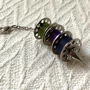 May include: A silver metal thread holder with a chain and a sharp point. It has three spools of thread, one green, one purple, and one blue.