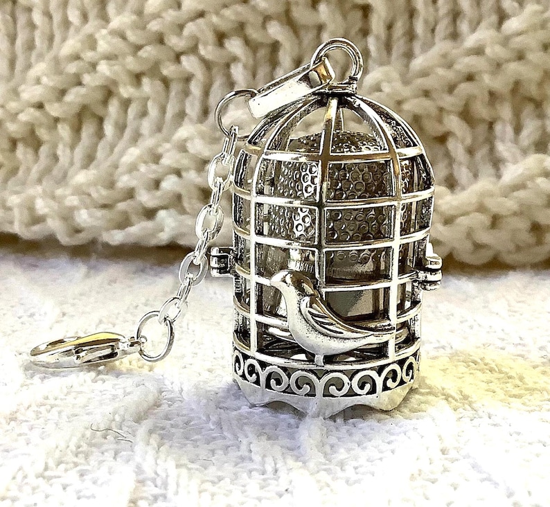 Silver tone thimble cage featuring a bird motif with a chain at the top and a bird clasp at the end. Hinged opening with space for a thimble inside.