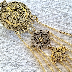 May include: Gold-toned body jewelry featuring an ornate circular pendant with bird and floral designs. The pendant is connected to a series of delicate chains and decorative elements, creating an elegant and detailed accessory.