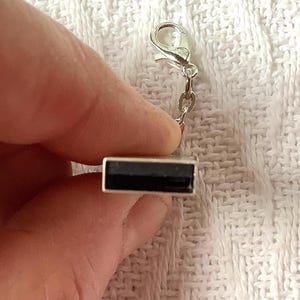 May include: A silver-toned rectangular charm with a black center, attached to a small chain and a clasp. The charm is held against a white, textured background. This jewelry accessory is suitable for bracelets or necklaces.