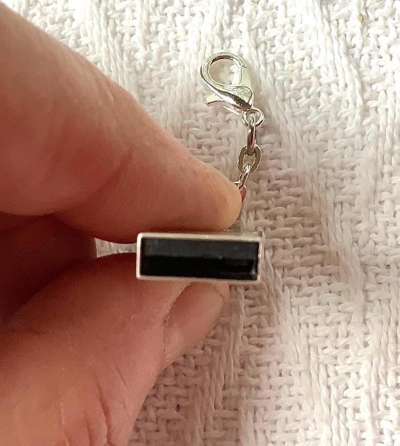 May include: A silver-toned rectangular charm with a black center, attached to a small chain and a clasp. The charm is held against a white, textured background. This jewelry accessory is suitable for bracelets or necklaces.