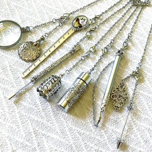 May include: A collection of silver-toned metal accessories attached to chains, including a magnifying glass, a floral embossed disc, a ruler, a birdcage charm, a cylindrical container, a slender pen, and a pointed tool with a decorative filigree charm.
